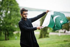 l'homme pakistano bangladesh porte des vêtements traditionnels noirs et des lunettes pose à l'extérieur, tenant le drapeau du pakistan. photo