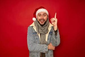 l'homme souriant excité au chapeau de noël tient l'index vers le haut se réjouit de la bonne idée de cadeau pour le nouvel an. concept de fond de studio rouge isolé - remue-méninges, vacances, promotions, remises, cadeaux, vente photo