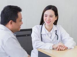 belle jeune femme médecin asiatique professionnelle parlant avec un homme patient de sa douleur et de ses symptômes pendant qu'ils mettent un masque facial et un thermomètre sur la table dans la salle d'examen photo