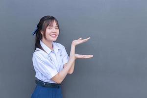 jolie lycéenne asiatique en uniforme scolaire avec des sourires confiants pendant qu'elle regarde la caméra pour présenter quelque chose de joyeux avec du gris en arrière-plan. photo