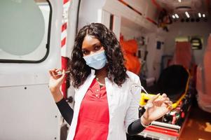 paramédic féminin afro-américain dans le masque médical de protection du visage debout devant la voiture d'ambulance. photo