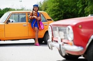 belle dame afro-américaine avec des sacs à provisions debout près d'une voiture rétro classique orange. photo