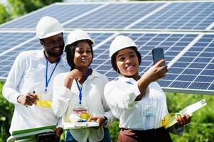 un technicien afro-américain vérifie l'entretien des panneaux solaires. groupe de trois ingénieurs noirs réunis à la station solaire. faire selfie par téléphone. photo