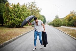 grand homme à la barbe arabe à la mode portant une chemise, un jean et des lunettes de soleil marchant au parc avec un parapluie et un manteau à portée de main. photo