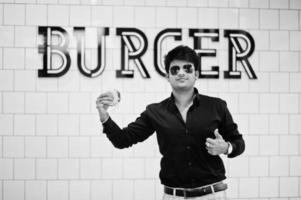 homme indien élégant à lunettes de soleil au café de restauration rapide avec hamburger à portée de main contre le signe de burger sur le mur. photo
