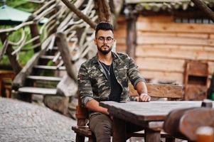 impressionnant beau grand homme macho à barbe arabe dans des verres et une veste militaire assis à une table en bois extérieure du restaurant. photo