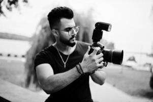 impressionnant beau grand photographe macho à barbe arabe dans des verres et un t-shirt noir avec un appareil photo professionnel à portée de main.