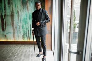 élégant gentleman afro-américain dans une élégante veste noire, tenant un bâton de marche rétro comme flacon de canne ou canne basculante avec poignée en boule de diamant doré. riche homme afro à la mode. photo