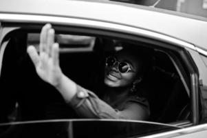 riche femme africaine d'affaires sur des lunettes de soleil s'asseoir à la voiture suv et montrer la main de la fenêtre. photo