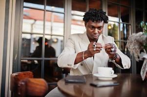 homme afro élégant en costume beige old school assis sur un café avec une tasse de café. jeune homme africain à la mode en veste décontractée sur torse nu. photo