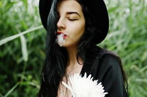 sensuelle fille fumeuse tout en noir, lèvres rouges et chapeau. une femme dramatique gothique tient une fleur de chrysanthème blanche et fume sur un roseau commun. photo