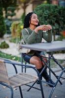 élégantes femmes afro-américaines à la mode en pull vert et jupe noire ont posé un café en plein air, assis à table avec une tasse de café.