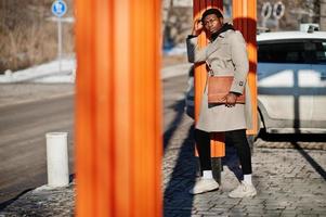 bel homme afro-américain posant à l'extérieur avec un chapeau noir et un manteau beige avec un dossier à la main. photo