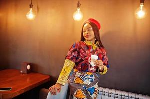 femme afro-américaine enthousiaste dans une tenue colorée à la mode avec un béret rouge qui se refroidit dans un café confortable avec une tasse de latte chaud dans les mains. photo