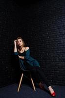 belle fille brune porte une veste noire et verte en talons hauts rouges, assise et posant sur une chaise au studio contre un mur de briques sombres. portrait de modèle de studio. photo