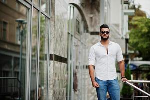 grand modèle d'homme arabe élégant en chemise blanche, jeans et lunettes de soleil posés dans la rue de la ville. Barbe mec arabe attrayant. photo
