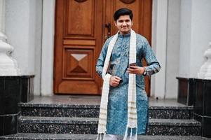 l'homme indien porte des vêtements traditionnels avec un foulard blanc posé à l'extérieur contre la porte du bâtiment avec un téléphone portable à portée de main. photo