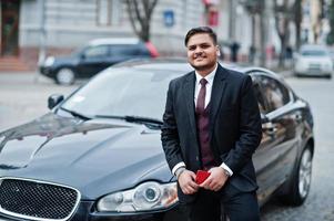 homme d'affaires indien élégant en tenue de soirée avec téléphone portable debout contre une voiture d'affaires noire dans la rue de la ville. photo