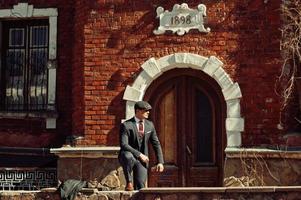 portrait d'un homme d'affaires arabe anglais rétro des années 1920 portant un costume sombre, une cravate et une casquette plate debout contre une vieille maison en brique année 1898. photo
