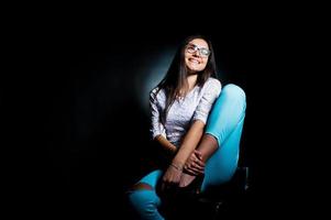 portrait d'une jolie jeune femme en haut blanc et pantalon bleu assis posant avec ses lunettes dans le noir. photo