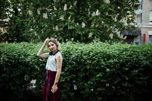 fille modèle blonde à la mode et belle en élégante jupe en velours rouge, chemisier blanc, posée au parc avec téléphone et écouteurs. photo