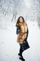 élégance bouclée fille en manteau de fourrure et sac à main au parc forestier enneigé en hiver. photo