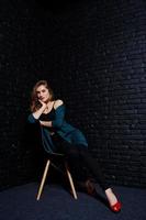 belle fille brune porte une veste noire et verte en talons hauts rouges, assise et posant sur une chaise au studio contre un mur de briques sombres. portrait de modèle de studio. photo