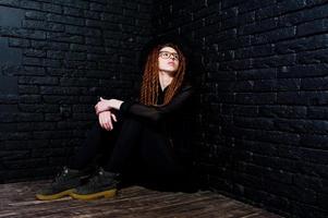 tournage en studio d'une fille en noir avec des dreads, des lunettes et un chapeau sur fond de briques. photo