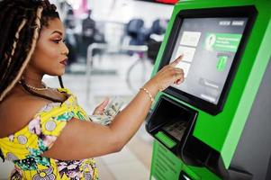 jolie fille afro-américaine de petite taille avec des dreadlocks, porter une robe jaune colorée, retirer de l'argent de la carte de crédit au guichet automatique. photo