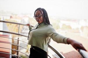 portrait de la ville d'une jeune femme positive à la peau foncée portant un sweat à capuche vert et des lunettes debout au balkony. photo