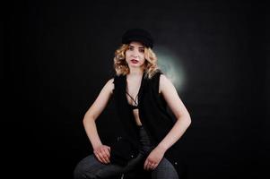 portrait en studio d'une fille blonde en vêtements noirs, soutien-gorge et casquette sur fond sombre. photo