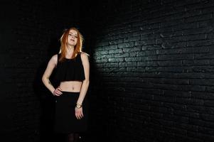 portrait d'une belle fille rousse en haut noir et jupe noire posant dans le studio à côté du mur de briques. photo