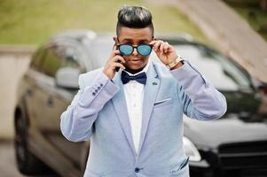 homme arabe élégant en veste, noeud papillon et lunettes de soleil contre une voiture suv noire. riche homme d'affaires arabe parlant sur téléphone mobile. photo