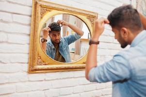 portrait d'un beau sud-asiatique barbu réussi, jeune pigiste indien en chemise de jeans bleu debout dans un café et regarde le miroir. photo