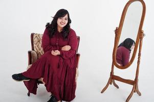 jolie femme sud-asiatique en robe rouge foncé posée au studio sur fond blanc contre le miroir et assis à la chaise. photo