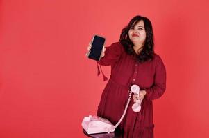 jolie femme sud-asiatique en robe rouge foncé posée au studio sur fond rose et utilise un vieux téléphone vintage avec un téléphone mobile moderne. photo