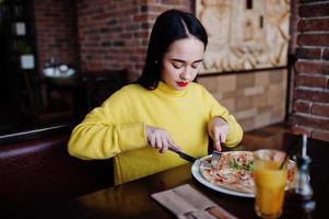 drôle de fille brune en pull jaune, manger de la pizza au restaurant. photo