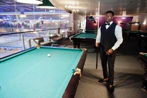 jeune bel homme africain vêtu d'une chemise blanche, d'un gilet noir et d'un nœud papillon jouant au billard. photo