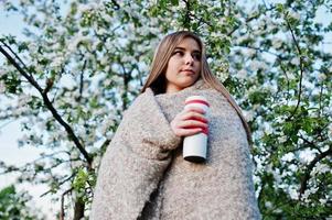 jeune fille brune sur plaid contre l'arbre de fleurs de printemps et tenant un thermos de thé à la main. photo