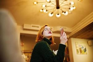 joyeuse jeune belle femme rousse à lunettes, pull en laine chaude verte, utilisant son téléphone, assise sur son lieu de travail au café. photo