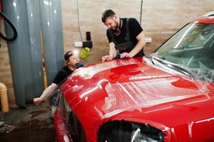 le travailleur du service automobile a mis un film anti-gravier sur une carrosserie de voiture rouge à l'atelier de détail du véhicule. protection de voiture avec des films spéciaux. photo