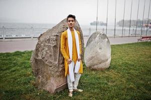 homme élégant indien en vêtements traditionnels jaunes avec foulard blanc posé en plein air contre de grosses pierres. photo