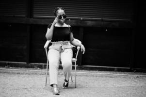 élégante femme afro-américaine à lunettes de soleil posée en plein air sur une chaise. photo