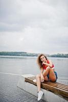 fille modèle bouclée sexy en haut rouge, short en jean jeans, lunettes et baskets posées sur un banc contre le lac. photo
