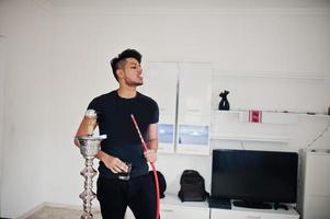 homme indien beau et à la mode en noir debout dans la chambre, fumant le narguilé avec des lunettes à portée de main. photo