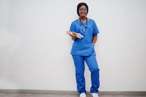 portrait d'une jeune femme médecin pédiatre afro-américaine heureuse en manteau uniforme bleu et stéthoscope avec des livres à portée de main. soins de santé, médecine, spécialiste de la médecine - concept. photo