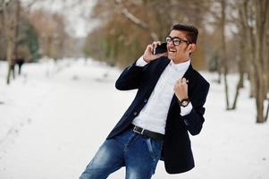 homme étudiant indien élégant en costume et lunettes posé à la journée d'hiver en plein air et parlant sur téléphone portable, montre des émotions heureuses. photo