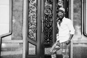 homme afro-américain élégant en chemise blanche et pantalon coloré avec chapeau et lunettes posés à l'extérieur. garçon modèle à la mode noir. photo