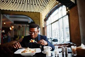 homme afro-américain à la mode en costume et lunettes assis au café et manger de la salade. photo