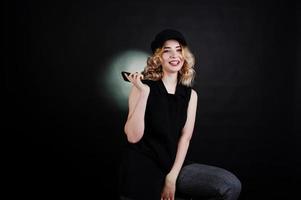 portrait en studio d'une fille blonde en vêtements noirs et casquette avec téléphone portable à portée de main sur fond sombre. photo